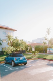A blue car is parked in an empty parking space under a clear, sunny sky. Palm trees and tropical vegetation surround a white building with a terracotta roof in the background.