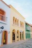 A row of colorful, Mediterranean-style buildings featuring arched doorways and windows. Each building is painted in a different pastel color, including light blue, yellow, and green. The architecture shows a combination of modern and traditional styles with ornate balconies and cobblestone pavement in front.