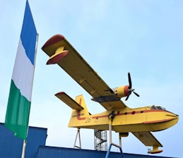 A large yellow amphibious airplane with red accents is mounted on a display stand. The airplane has propellers and is marked with the number 26 and the words 'Sécurité Civile' on the side. To the left of the aircraft, a tall flagpole displays a blue, white, and green flag. The sky is clear, and the scene includes parts of a blue building structure.