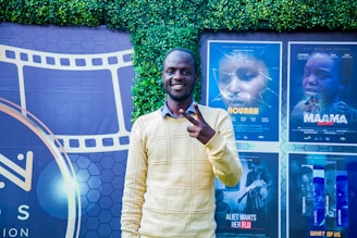 A person stands smiling and making a peace sign in front of a backdrop featuring movie posters and a film reel design. The background includes a green leafy wall and posters for different films in various shades.