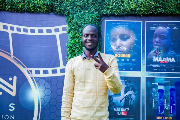 A person stands smiling and making a peace sign in front of a backdrop featuring movie posters and a film reel design. The background includes a green leafy wall and posters for different films in various shades.