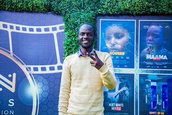 A person stands smiling and making a peace sign in front of a backdrop featuring movie posters and a film reel design. The background includes a green leafy wall and posters for different films in various shades.