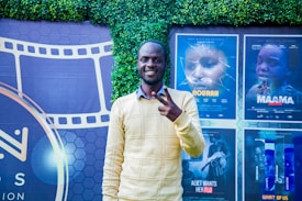 A person stands smiling and making a peace sign in front of a backdrop featuring movie posters and a film reel design. The background includes a green leafy wall and posters for different films in various shades.
