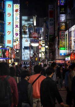 A bustling city street at night, illuminated by numerous colorful neon signs in various languages. A diverse crowd of people walk along the sidewalk, some wearing masks. The atmosphere is vibrant and energetic, typical of an urban nightlife scene.