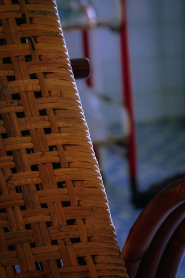 A close-up view of a wicker or rattan chair with intricate weaving. The background features blurred elements including a red vertical bar and a blue-toned floor.