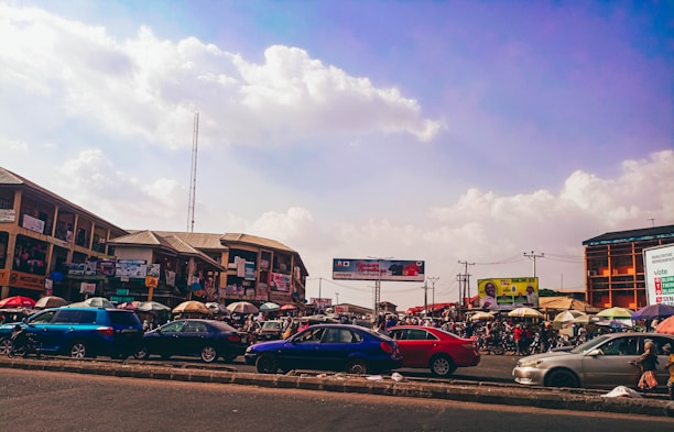 A bustling marketplace with numerous cars parked along the road. Stalls with colorful umbrellas line the street, and people are visible engaging in market activities. Buildings in the background display various signs and advertisements. The sky is partly cloudy, creating a vibrant urban scene.