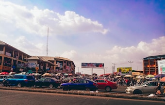A bustling marketplace with numerous cars parked along the road. Stalls with colorful umbrellas line the street, and people are visible engaging in market activities. Buildings in the background display various signs and advertisements. The sky is partly cloudy, creating a vibrant urban scene.