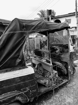 A dilapidated and abandoned three-wheeler vehicle is parked on a rough street. It appears old and deteriorated, with parts of the body and interior exposed. The surroundings include a residential boundary wall and some nearby structures, such as a house and utility poles.