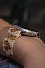 A close-up of a wrist wearing a smartwatch with a handmade beaded strap. The beads are arranged in a geometric pattern featuring white, gold, and orange colors.