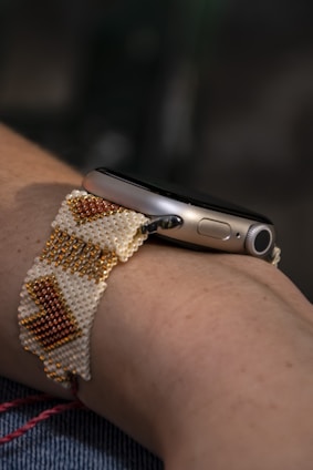 A close-up of a wrist wearing a smartwatch with a handmade beaded strap. The beads are arranged in a geometric pattern featuring white, gold, and orange colors.