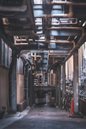 An urban alleyway is filled with industrial piping and ventilation systems. Large metal structures and various tubes are overhead. On the ground level, there are bicycles and an orange traffic cone. The scene is mostly shaded, with light filtering from above.