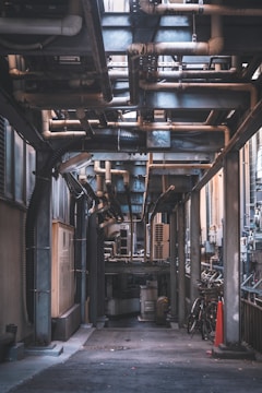 An urban alleyway is filled with industrial piping and ventilation systems. Large metal structures and various tubes are overhead. On the ground level, there are bicycles and an orange traffic cone. The scene is mostly shaded, with light filtering from above.