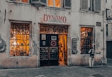 A vintage-looking bicycle shop with a weathered facade and a sign above the door reading 'Dynamo L'Atelier du Vélo.' The shop windows are filled with bicycle parts and equipment, and warm light emanates from inside, suggesting a cozy and inviting atmosphere. A person stands near one of the windows, appearing to be looking inside.