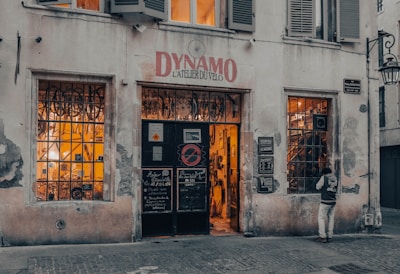 A vintage-looking bicycle shop with a weathered facade and a sign above the door reading 'Dynamo L'Atelier du Vélo.' The shop windows are filled with bicycle parts and equipment, and warm light emanates from inside, suggesting a cozy and inviting atmosphere. A person stands near one of the windows, appearing to be looking inside.