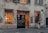 A vintage-looking bicycle shop with a weathered facade and a sign above the door reading 'Dynamo L'Atelier du Vélo.' The shop windows are filled with bicycle parts and equipment, and warm light emanates from inside, suggesting a cozy and inviting atmosphere. A person stands near one of the windows, appearing to be looking inside.