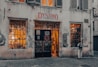 A vintage-looking bicycle shop with a weathered facade and a sign above the door reading 'Dynamo L'Atelier du V&eacute;lo.' The shop windows are filled with bicycle parts and equipment, and warm light emanates from inside, suggesting a cozy and inviting atmosphere. A person stands near one of the windows, appearing to be looking inside.