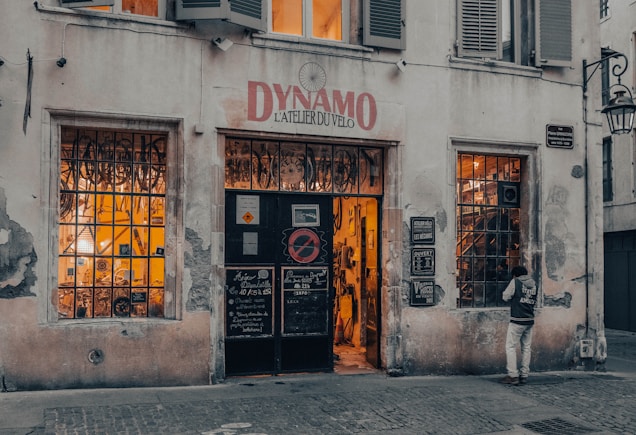 A vintage-looking bicycle shop with a weathered facade and a sign above the door reading 'Dynamo L'Atelier du V&eacute;lo.' The shop windows are filled with bicycle parts and equipment, and warm light emanates from inside, suggesting a cozy and inviting atmosphere. A person stands near one of the windows, appearing to be looking inside.