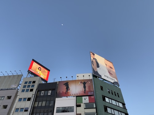 Multiple buildings with large billboards are seen against a clear blue sky. The billboards display various advertisements, including luxury fashion brand Prada and a vibrant ad featuring a person with Japanese text. There's a silhouette of a person on another sign, and a small white moon is visible in the sky.