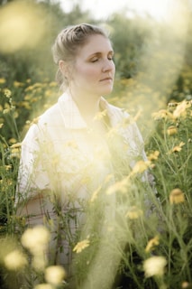 A person with closed eyes is surrounded by tall green plants and yellow flowers. The sunlight creates a soft, dreamy atmosphere with a gentle, natural ambiance.