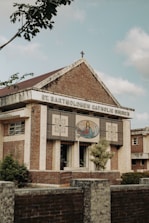 A brick and stone church building with a cross on the roof, featuring an artwork depicting a religious figure with open arms on the front facade. The name 'St. Bartholomew Catholic Church' is displayed prominently. Trees and shrubs are visible around the structure, and the sky appears partly cloudy.