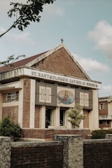 A brick and stone church building with a cross on the roof, featuring an artwork depicting a religious figure with open arms on the front facade. The name 'St. Bartholomew Catholic Church' is displayed prominently. Trees and shrubs are visible around the structure, and the sky appears partly cloudy.