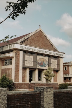 A brick and stone church building with a cross on the roof, featuring an artwork depicting a religious figure with open arms on the front facade. The name 'St. Bartholomew Catholic Church' is displayed prominently. Trees and shrubs are visible around the structure, and the sky appears partly cloudy.