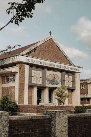 A brick and stone church building with a cross on the roof, featuring an artwork depicting a religious figure with open arms on the front facade. The name 'St. Bartholomew Catholic Church' is displayed prominently. Trees and shrubs are visible around the structure, and the sky appears partly cloudy.