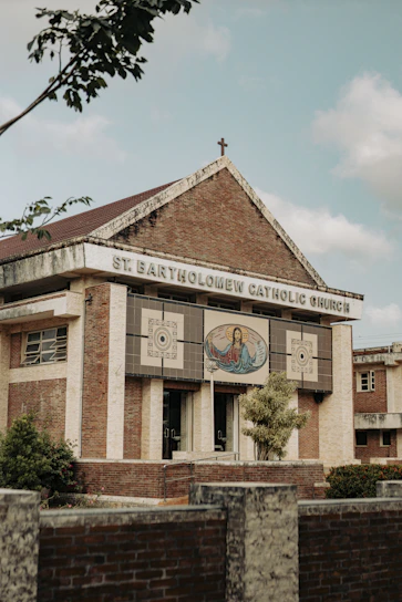 A brick and stone church building with a cross on the roof, featuring an artwork depicting a religious figure with open arms on the front facade. The name 'St. Bartholomew Catholic Church' is displayed prominently. Trees and shrubs are visible around the structure, and the sky appears partly cloudy.