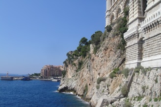 Large stone building set atop a rocky cliff with greenery. In the background, a harbor area with yachts and a breakwater is visible, along with residential buildings further in the distance. The sea is a vibrant blue, contrasting with the brown and green hues of the cliff.