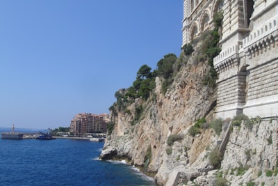 Large stone building set atop a rocky cliff with greenery. In the background, a harbor area with yachts and a breakwater is visible, along with residential buildings further in the distance. The sea is a vibrant blue, contrasting with the brown and green hues of the cliff.