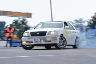 A white car is drifting around a corner on a track, creating smoke from the tires. The car has visible wear and tear, while a person in an orange safety vest is standing in the background, watching the event. There is a blurred audience in the backdrop.