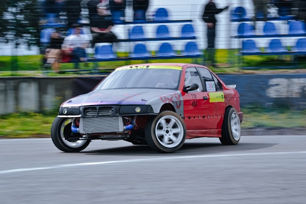 A red modified race car is drifting on a racetrack, tilting at an angle as it takes a corner. The car is equipped with a front-mounted intercooler and has large, white racing wheels. Spectators are visible in the background, sitting in blue seats behind a fence.