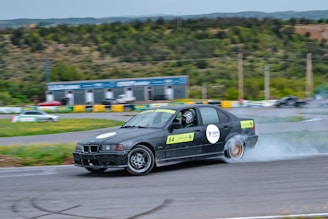 A black car with numbered signs is drifting on a racetrack, producing smoke from its rear tires. The driver is wearing a helmet. The surroundings include green hills, trees, and a small structure in the background.