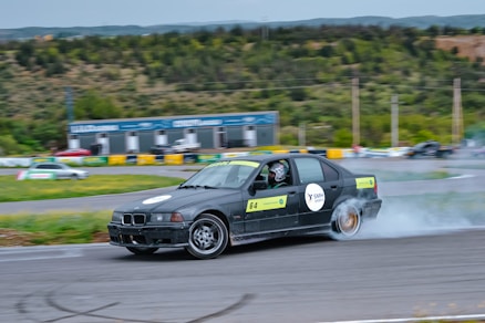 A black car with numbered signs is drifting on a racetrack, producing smoke from its rear tires. The driver is wearing a helmet. The surroundings include green hills, trees, and a small structure in the background.