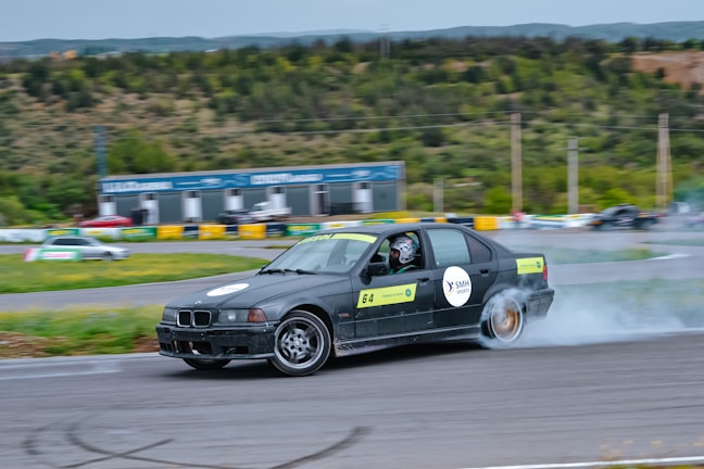 A black car with numbered signs is drifting on a racetrack, producing smoke from its rear tires. The driver is wearing a helmet. The surroundings include green hills, trees, and a small structure in the background.