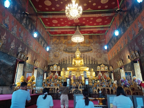A richly decorated temple interior with a large golden Buddha statue at the center, surrounded by ornate floral arrangements and traditional Thai artwork on the walls. There is a chandelier overhead and several people sitting on the floor in front of the altar, appearing to be in prayer or meditation.