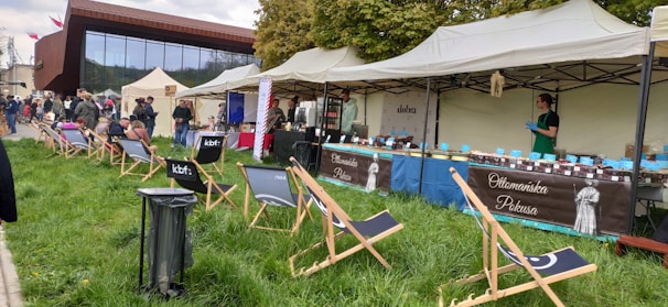 An outdoor market scene with several stalls set up under tents. A vendor wearing gloves is standing at a stall labeled 'Ottomańska Pokusa,' which is displaying various jars. There are several folding chairs on the grass, with people sitting and walking around in the background. The atmosphere appears casual and relaxed, with a mix of grassy area and modern building backdrop.