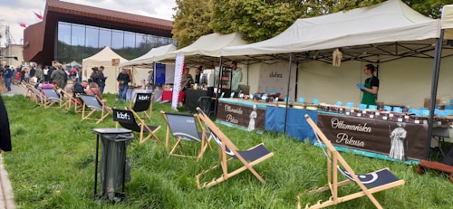 An outdoor market scene with several stalls set up under tents. A vendor wearing gloves is standing at a stall labeled 'Ottomańska Pokusa,' which is displaying various jars. There are several folding chairs on the grass, with people sitting and walking around in the background. The atmosphere appears casual and relaxed, with a mix of grassy area and modern building backdrop.