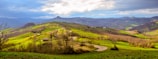 Rolling hills stretch across a vast landscape with vibrant green grass and scattered trees. A winding road cuts through the scene, leading towards distant hills and mountains under a partly cloudy sky.