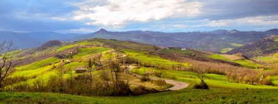 Rolling hills stretch across a vast landscape with vibrant green grass and scattered trees. A winding road cuts through the scene, leading towards distant hills and mountains under a partly cloudy sky.