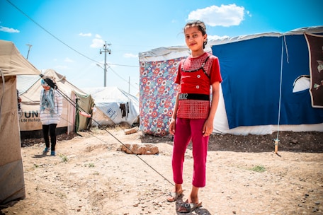 A young girl stands confidently in front of a tent decorated with a colorful floral sheet. The scene appears to be a refugee camp, as indicated by the UNHCR logo on nearby tents. Another person is visible in the background, partially obscured, wearing a headscarf and striped shirt. The ground is dry and rocky, and the sky is bright and clear with few clouds.