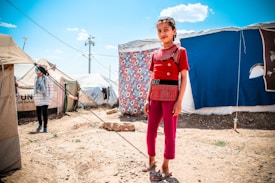A young girl stands confidently in front of a tent decorated with a colorful floral sheet. The scene appears to be a refugee camp, as indicated by the UNHCR logo on nearby tents. Another person is visible in the background, partially obscured, wearing a headscarf and striped shirt. The ground is dry and rocky, and the sky is bright and clear with few clouds.