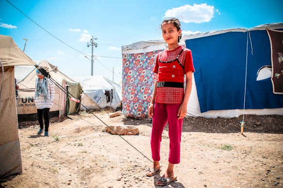 A young girl stands confidently in front of a tent decorated with a colorful floral sheet. The scene appears to be a refugee camp, as indicated by the UNHCR logo on nearby tents. Another person is visible in the background, partially obscured, wearing a headscarf and striped shirt. The ground is dry and rocky, and the sky is bright and clear with few clouds.