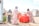 Four women and a young girl are engaged in daily activities near large red water containers. They are standing on a rocky, dusty ground with makeshift shelters in the background displaying UNHCR logos. The women are dressed in colorful headscarves and patterned dresses. One woman is carrying a box, while another is standing beside the containers.