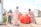 Four women and a young girl are engaged in daily activities near large red water containers. They are standing on a rocky, dusty ground with makeshift shelters in the background displaying UNHCR logos. The women are dressed in colorful headscarves and patterned dresses. One woman is carrying a box, while another is standing beside the containers.