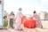 Four women and a young girl are engaged in daily activities near large red water containers. They are standing on a rocky, dusty ground with makeshift shelters in the background displaying UNHCR logos. The women are dressed in colorful headscarves and patterned dresses. One woman is carrying a box, while another is standing beside the containers.