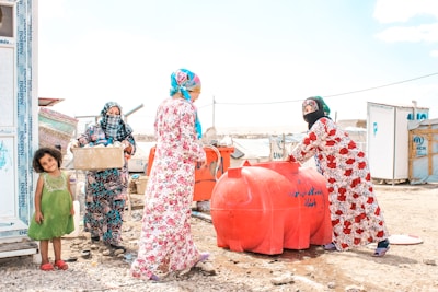Four women and a young girl are engaged in daily activities near large red water containers. They are standing on a rocky, dusty ground with makeshift shelters in the background displaying UNHCR logos. The women are dressed in colorful headscarves and patterned dresses. One woman is carrying a box, while another is standing beside the containers.