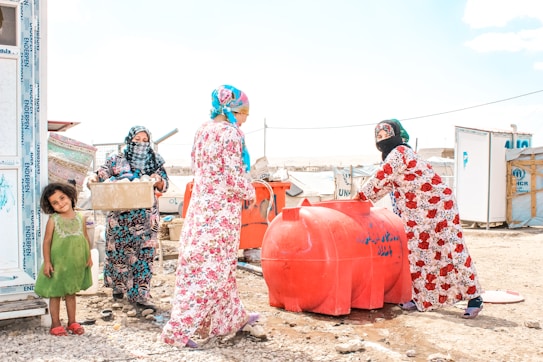 Four women and a young girl are engaged in daily activities near large red water containers. They are standing on a rocky, dusty ground with makeshift shelters in the background displaying UNHCR logos. The women are dressed in colorful headscarves and patterned dresses. One woman is carrying a box, while another is standing beside the containers.