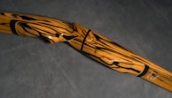 A wooden archery bow with a striking pattern of black swirls and lines against a light brown surface. The bow has a smooth finish and a curved design, set against a dark gray background.