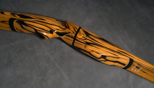 A wooden archery bow with a striking pattern of black swirls and lines against a light brown surface. The bow has a smooth finish and a curved design, set against a dark gray background.